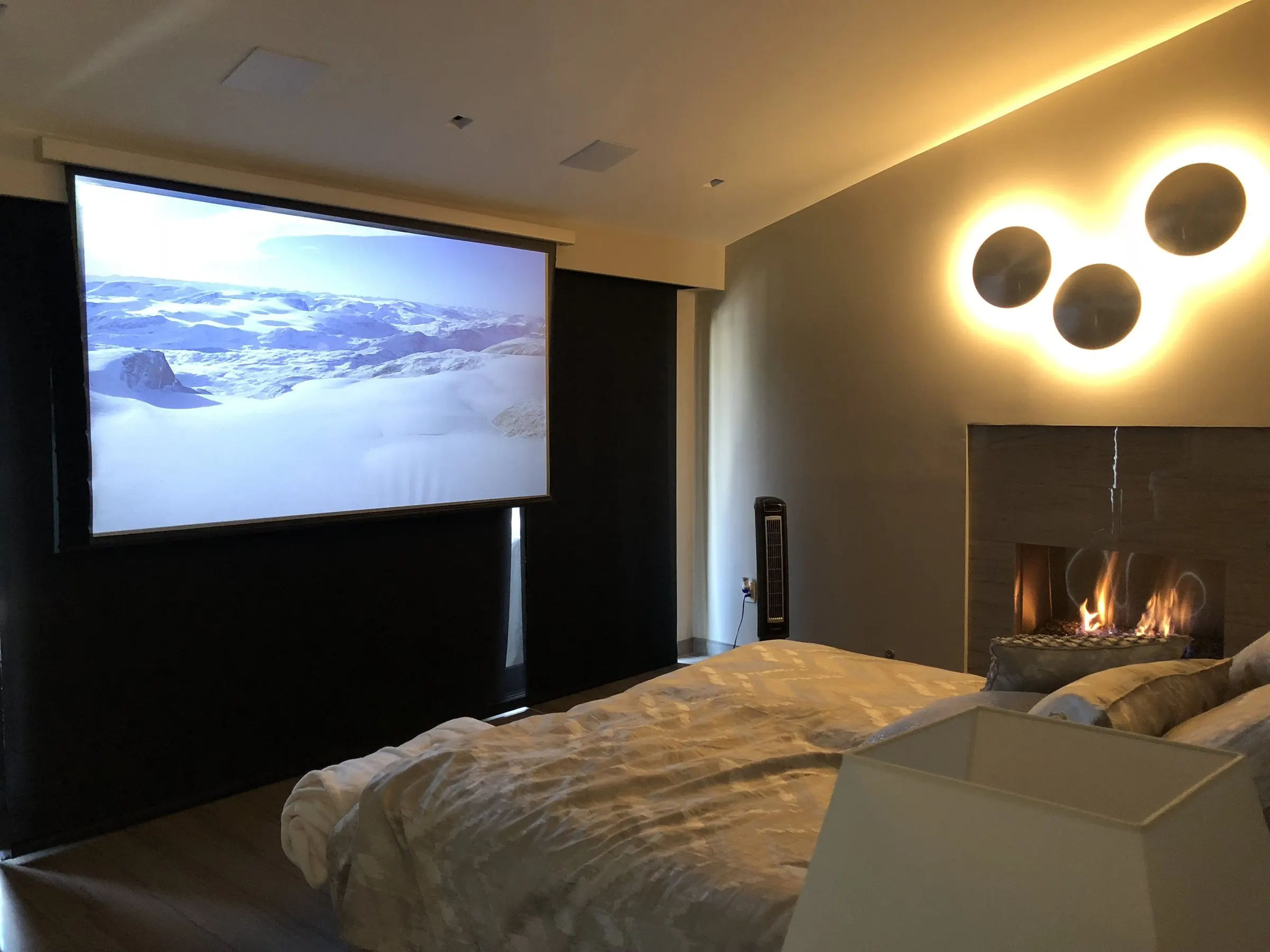 Bedroom with a projection screen lowered beside a fireplace and circular backlit wall sculptures