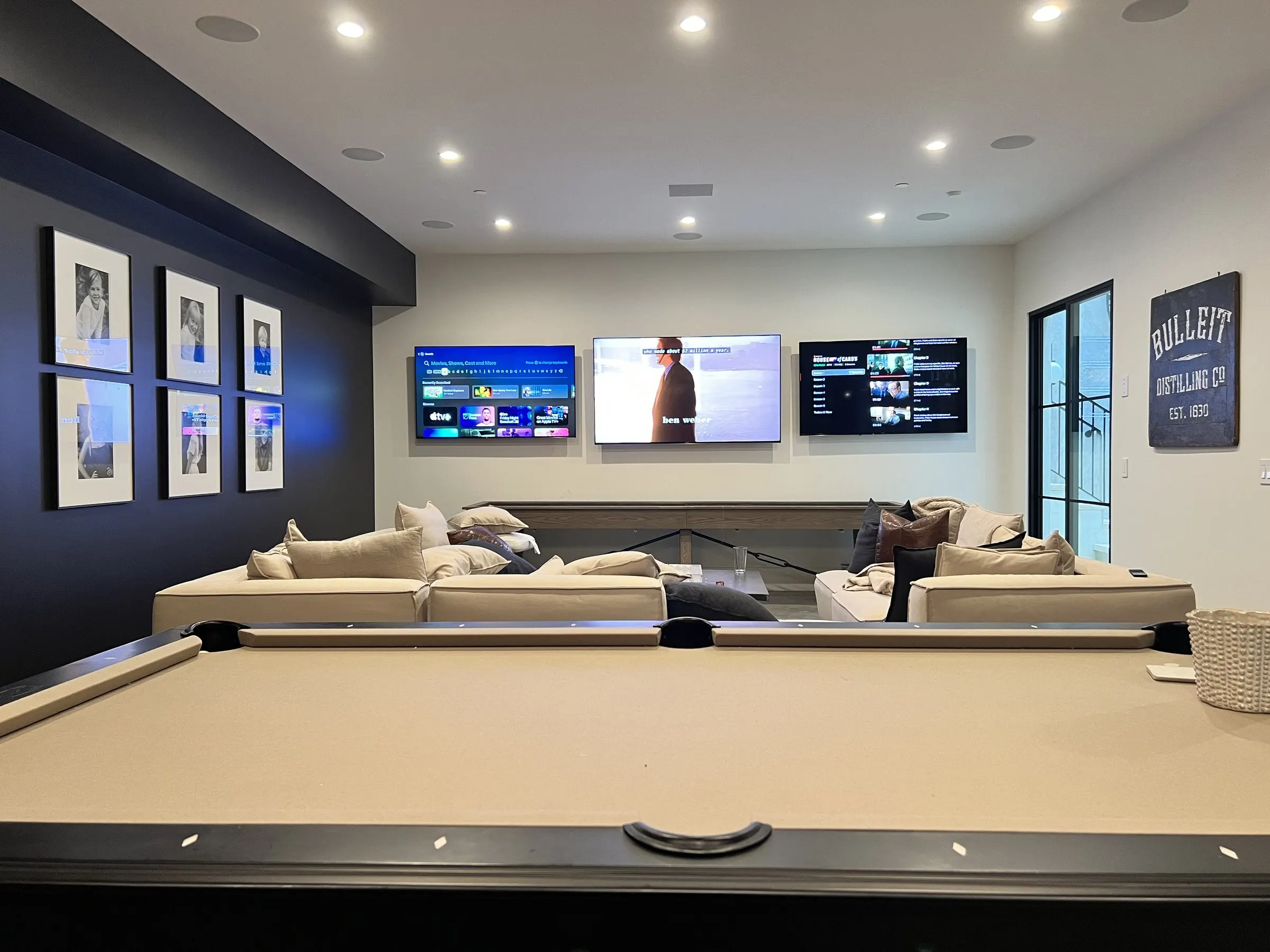 Game room with three wall-mounted displays above a sectional and a pool table in the foreground