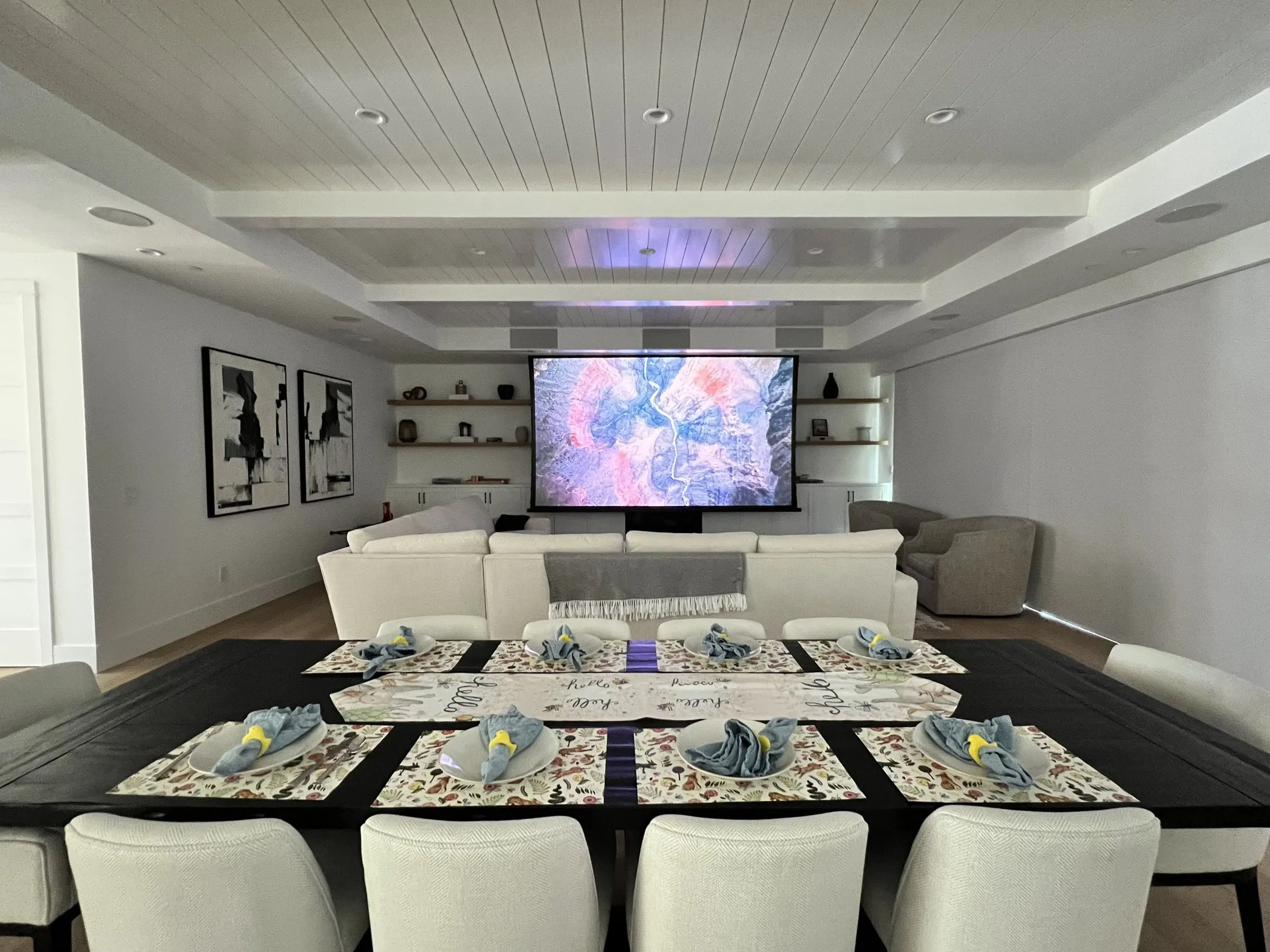 Open great room with a dining table in the foreground and a motorized screen lowered at the far end