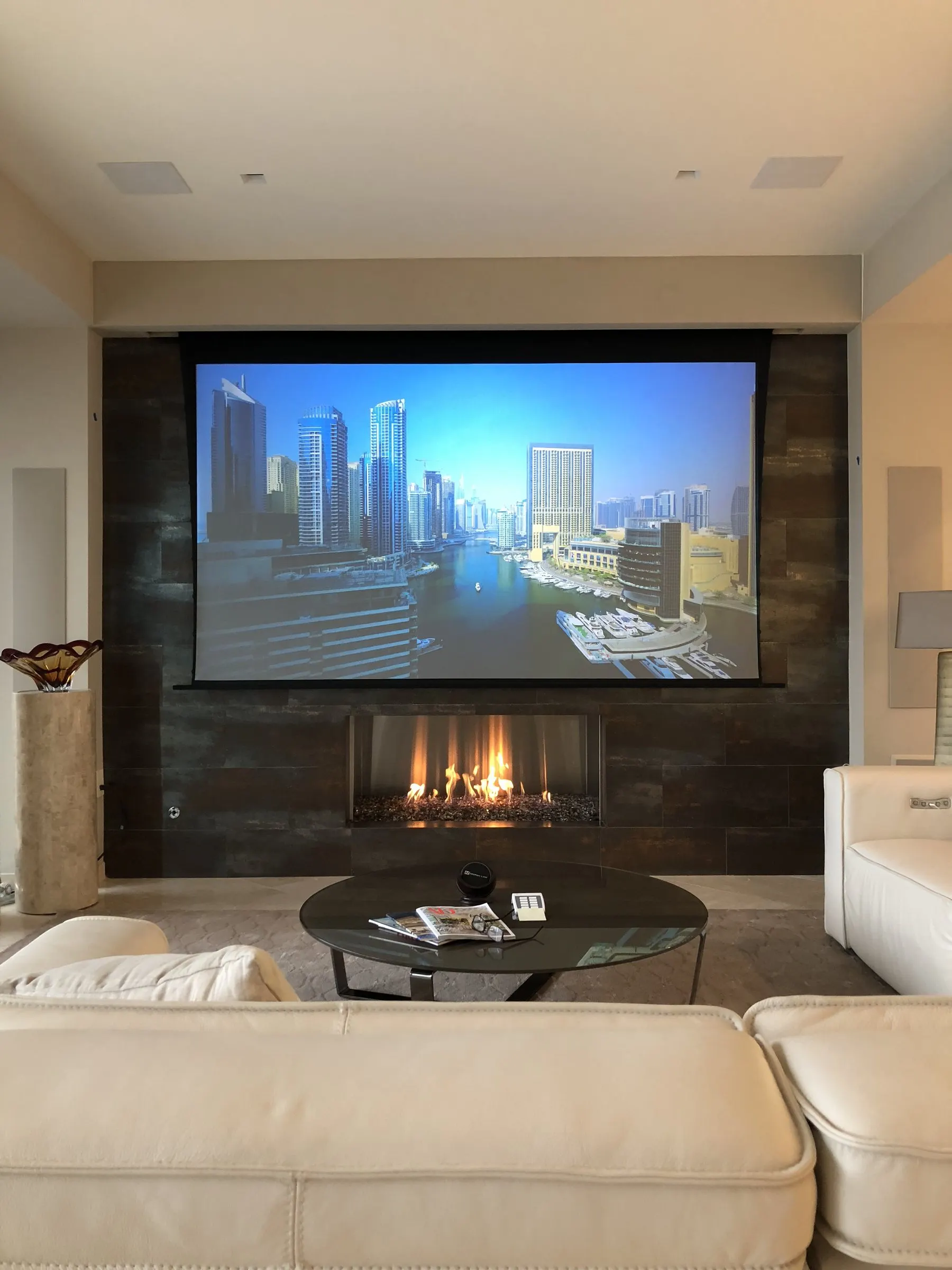 Living room with a motorized projection screen lowered above a lit linear fireplace, cream leather sofa