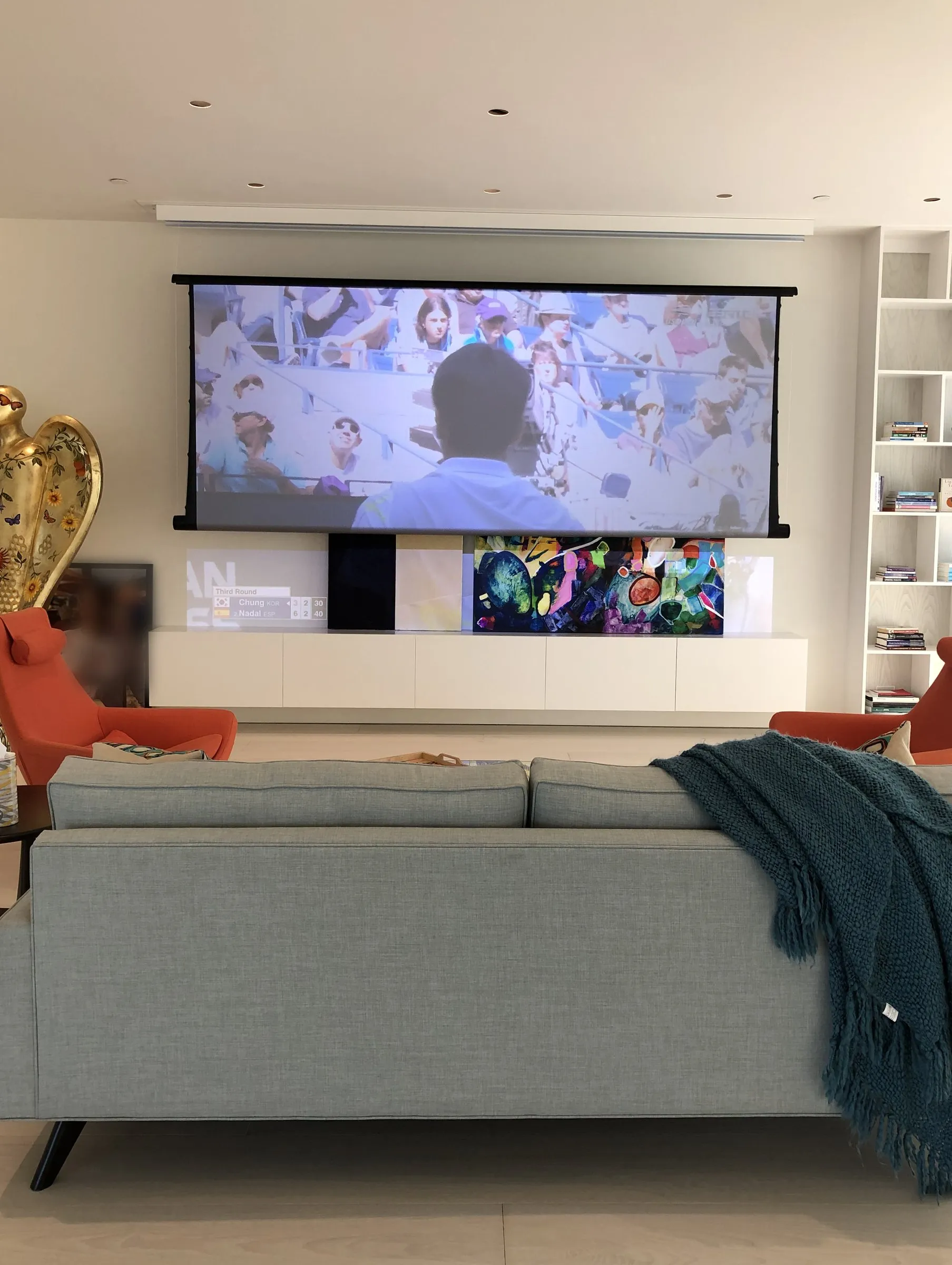 The same living room with a motorized projection screen lowered in front of the art wall, a film playing