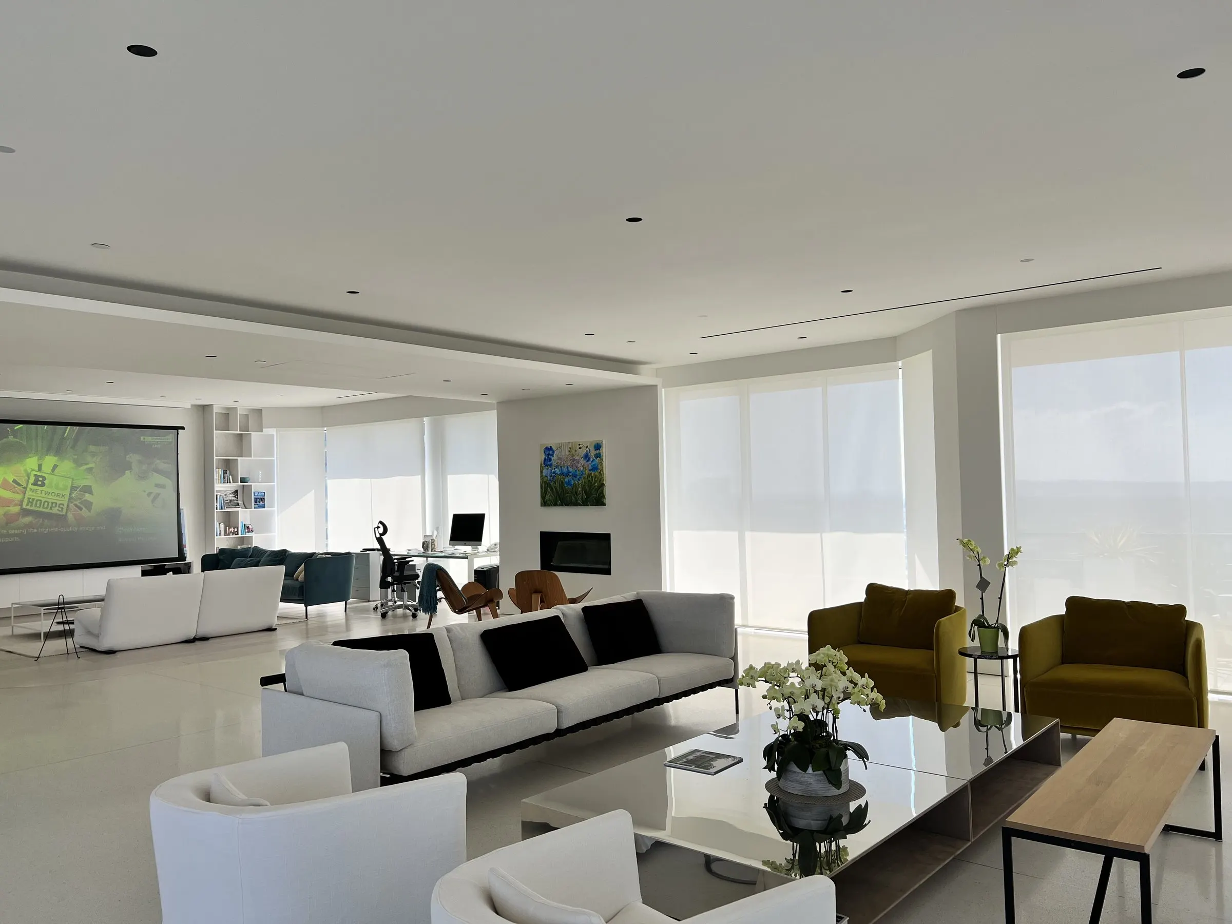 Wide open-plan living area with white motorized shades lowered and a projection screen at the far end