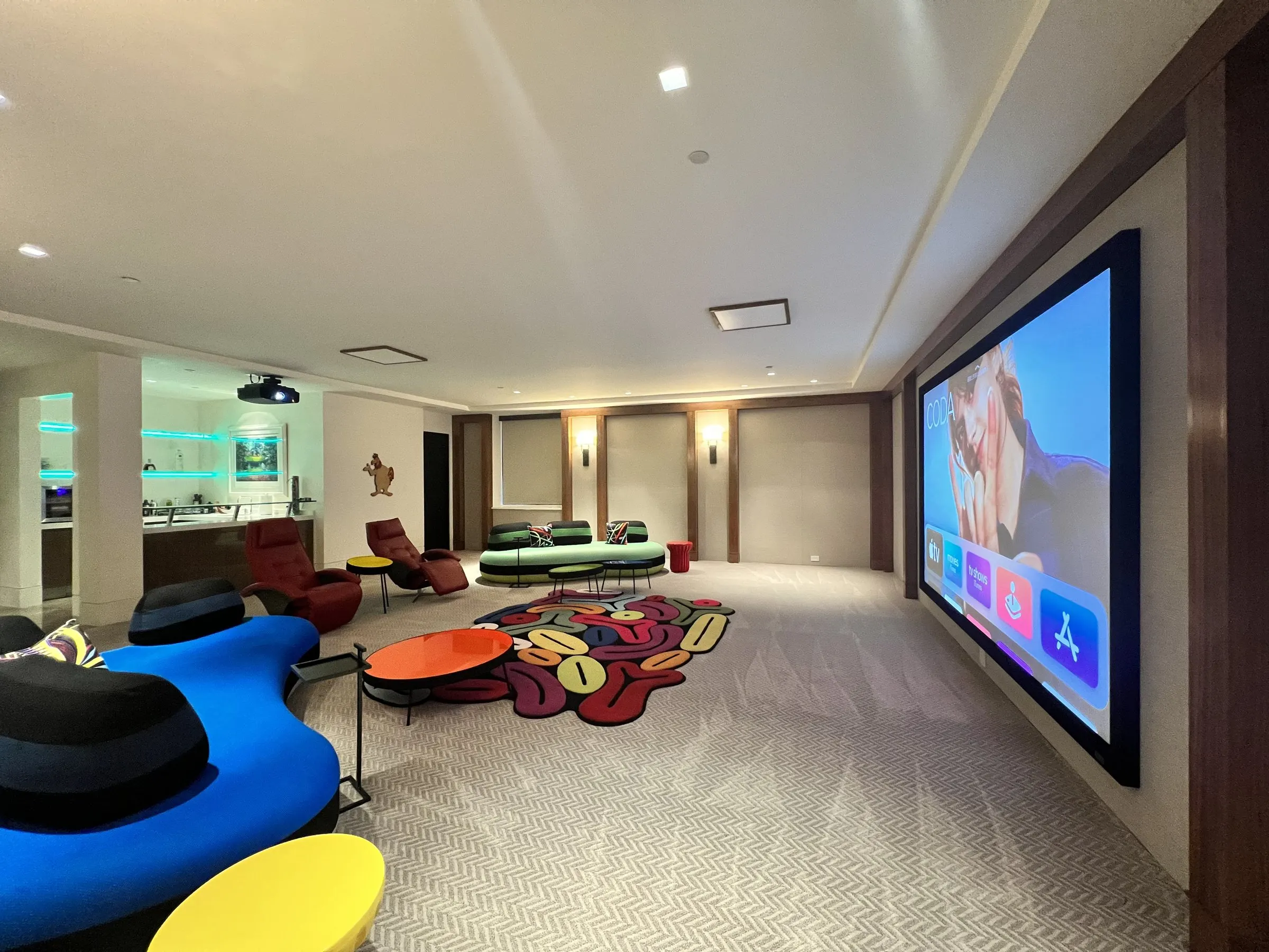 Long lounge theater with colorful modern seating, patterned rug, and a large wall-mounted projection screen