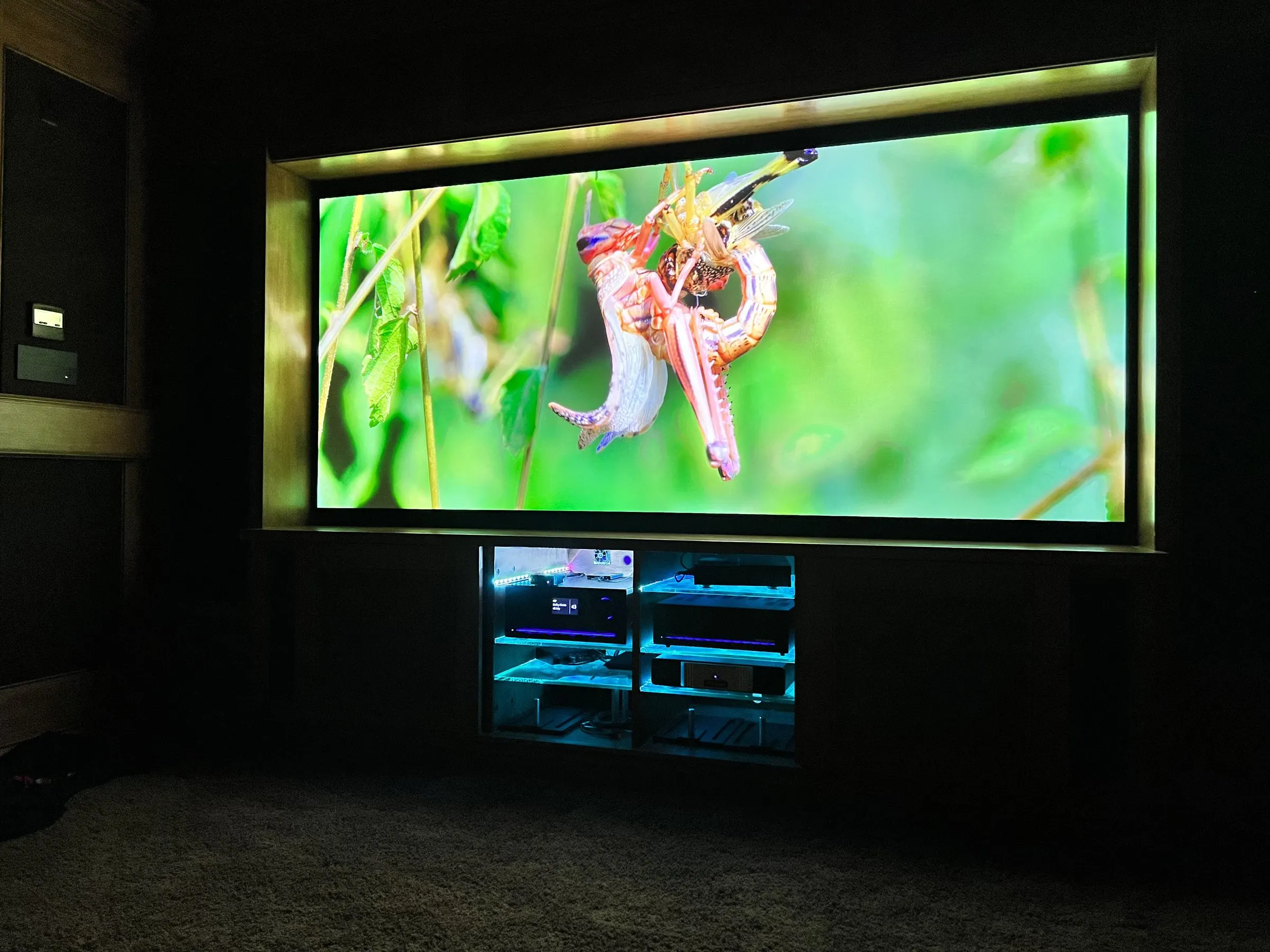 Dark home theater with a large illuminated projection screen framed in a lit reveal, equipment in a glass-front cabinet below