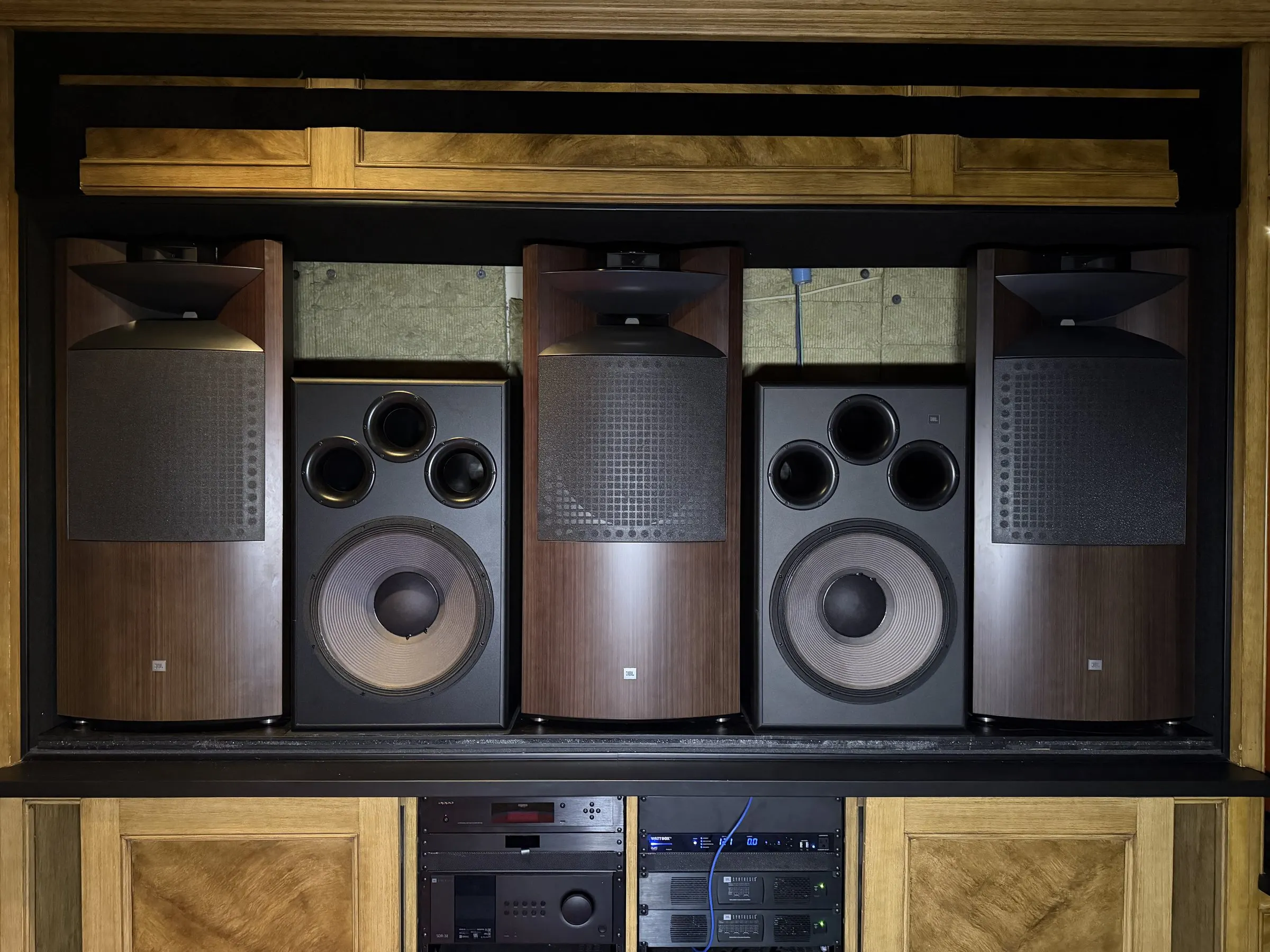 Behind-screen speaker wall with three large horn-loaded speakers and two subwoofers above an equipment rack