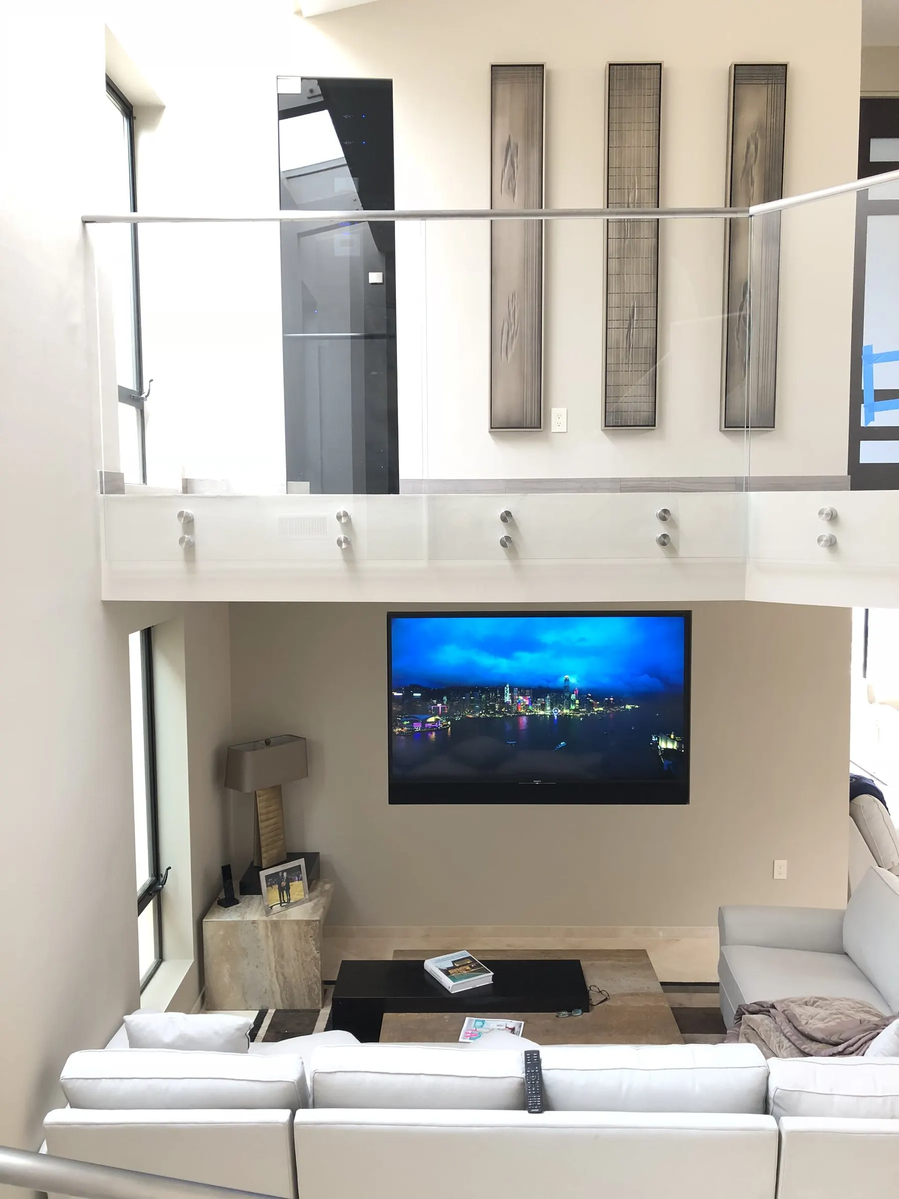 Television on the wall of a double-height room beneath a glass balcony